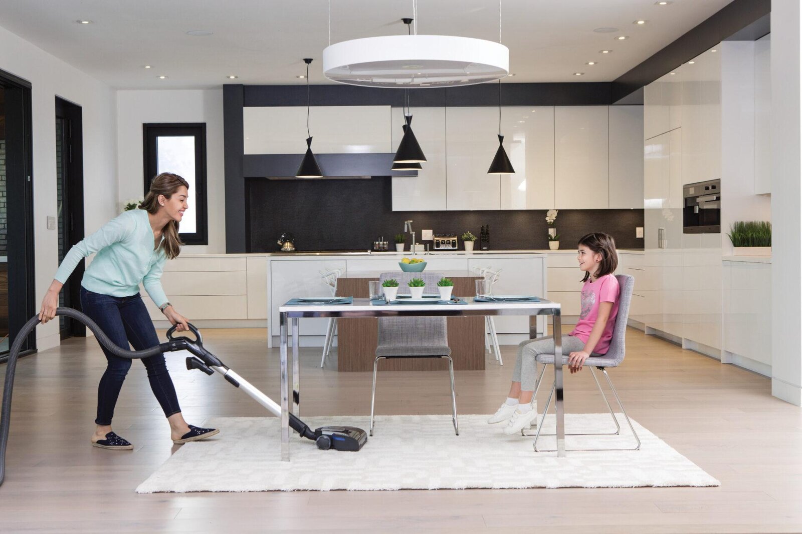Mom vacuuming a modern kitchen with a BEAM central vacuum while her daughter sits at the table